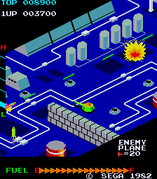 Zaxxon attract mode: an isometric view of a blue enemy fortress, its grey brick wall running diagonally across the middle of the screen with gun turrets, fuel tanks and radar dishes on the surface and a yellow explosion at the upper right, TOP 008900 and 1UP 003700 along the top, the ENEMY PLANE =20 scoring line at the lower right, and a green FUEL label beside a bar of yellow and red chevrons above the SEGA 1982 copyright line